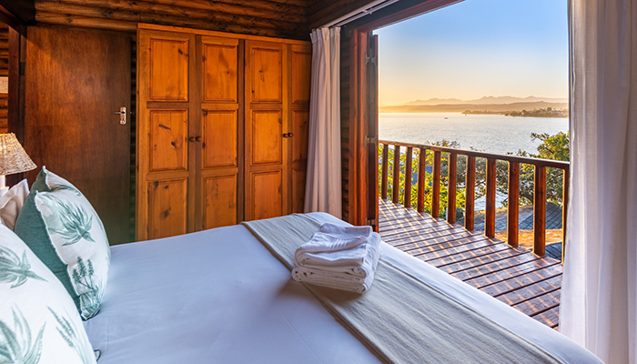 Timber chalet bedroom with open doors overlooking the Knysna lagoon at sunrise