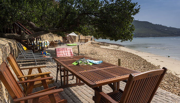 Wooden outdoor furniture on a shaded patio beside the Knysna lagoon at The Milkwood Collection