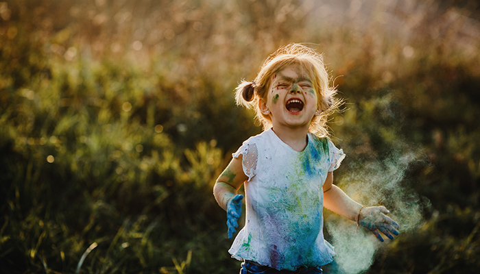 Child laughing outdoors with colourful paint on face and clothes during a fun family activity